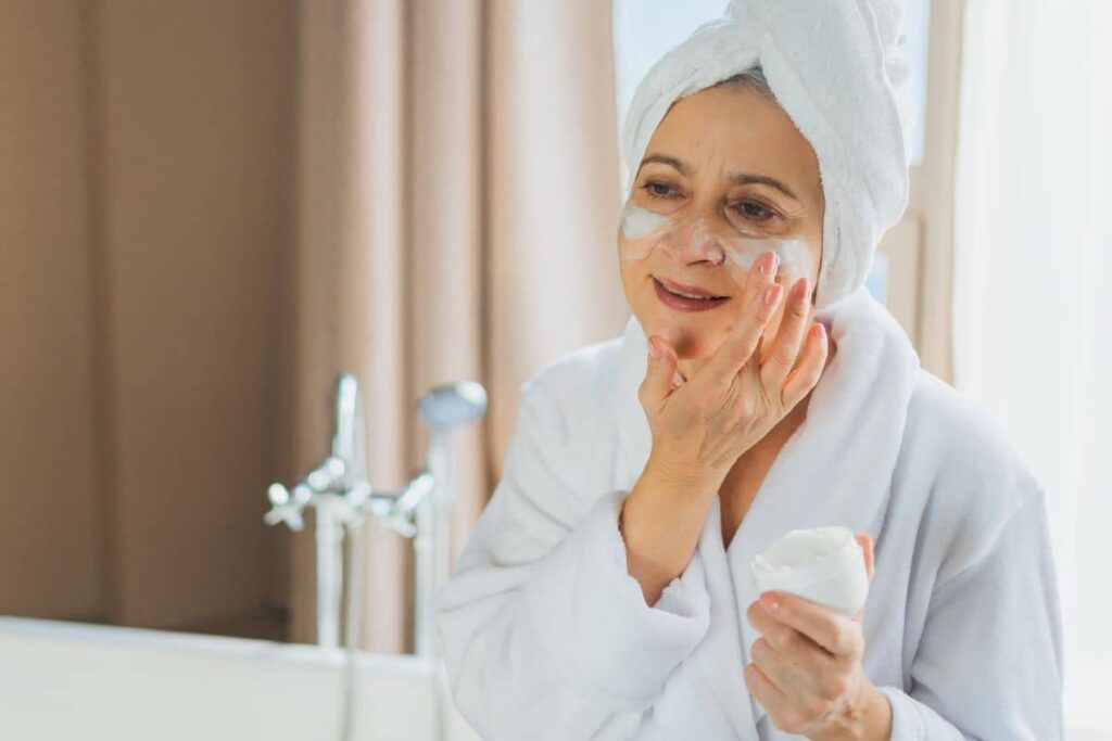 Older woman in bathrobe and towel applying moisturizer to face as part of facelift skincare preparation routine.