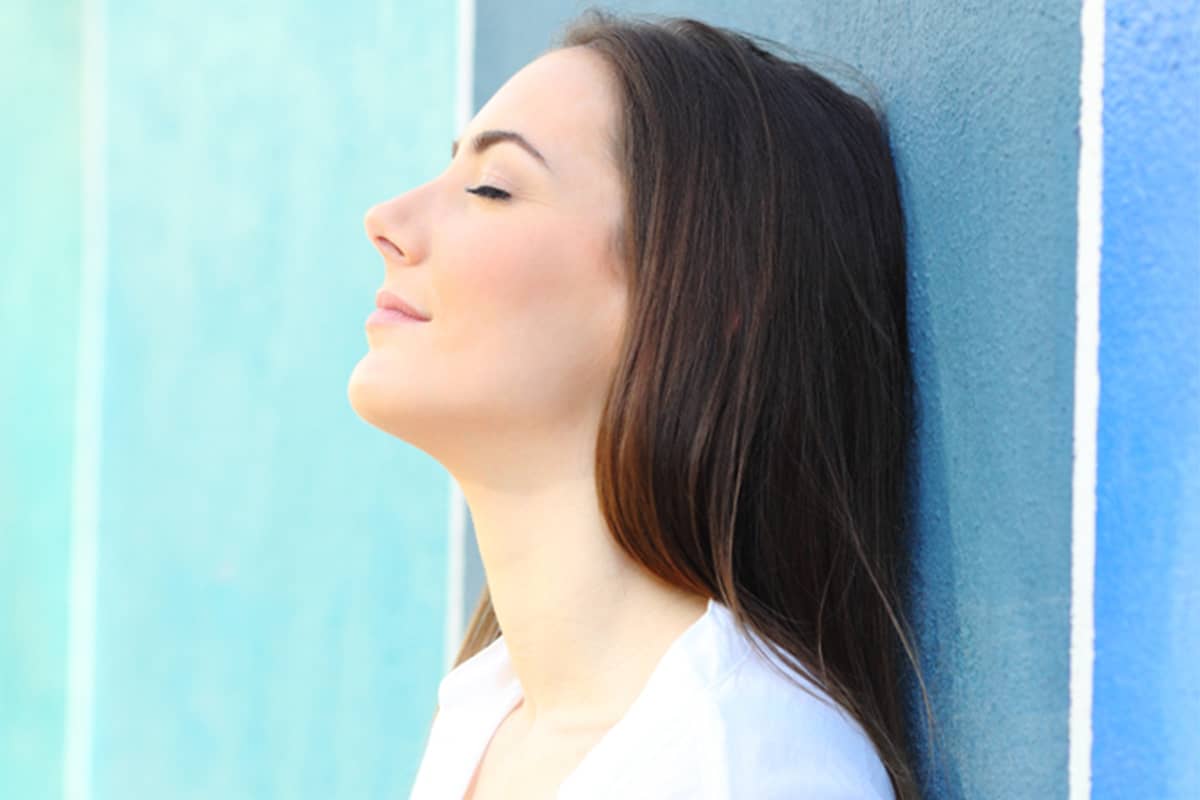 Side profile of a woman with a straight nasal bridge leaning against a blue wall with her eyes closed, representing the successful results of both functional and cosmetic rhinoplasty.