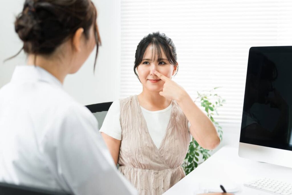 An Asian woman pointing to her nose during a consultation with a facial plastic surgeon to discuss her specific aesthetic goals.