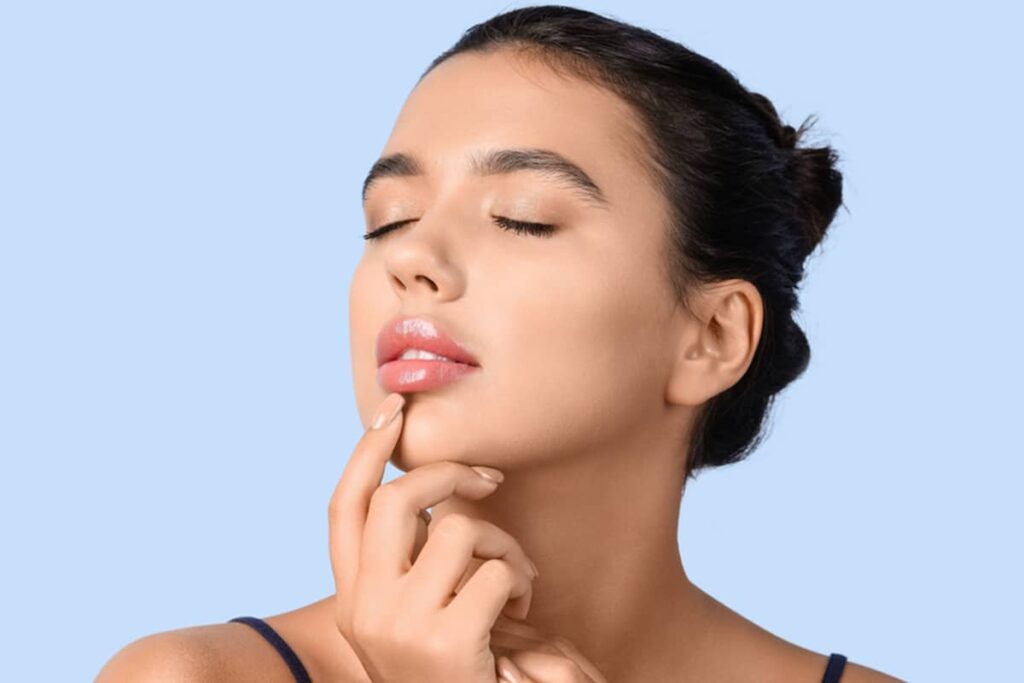 A woman with her eyes closed and hair in a bun gently touching her upper lip against a light blue background.