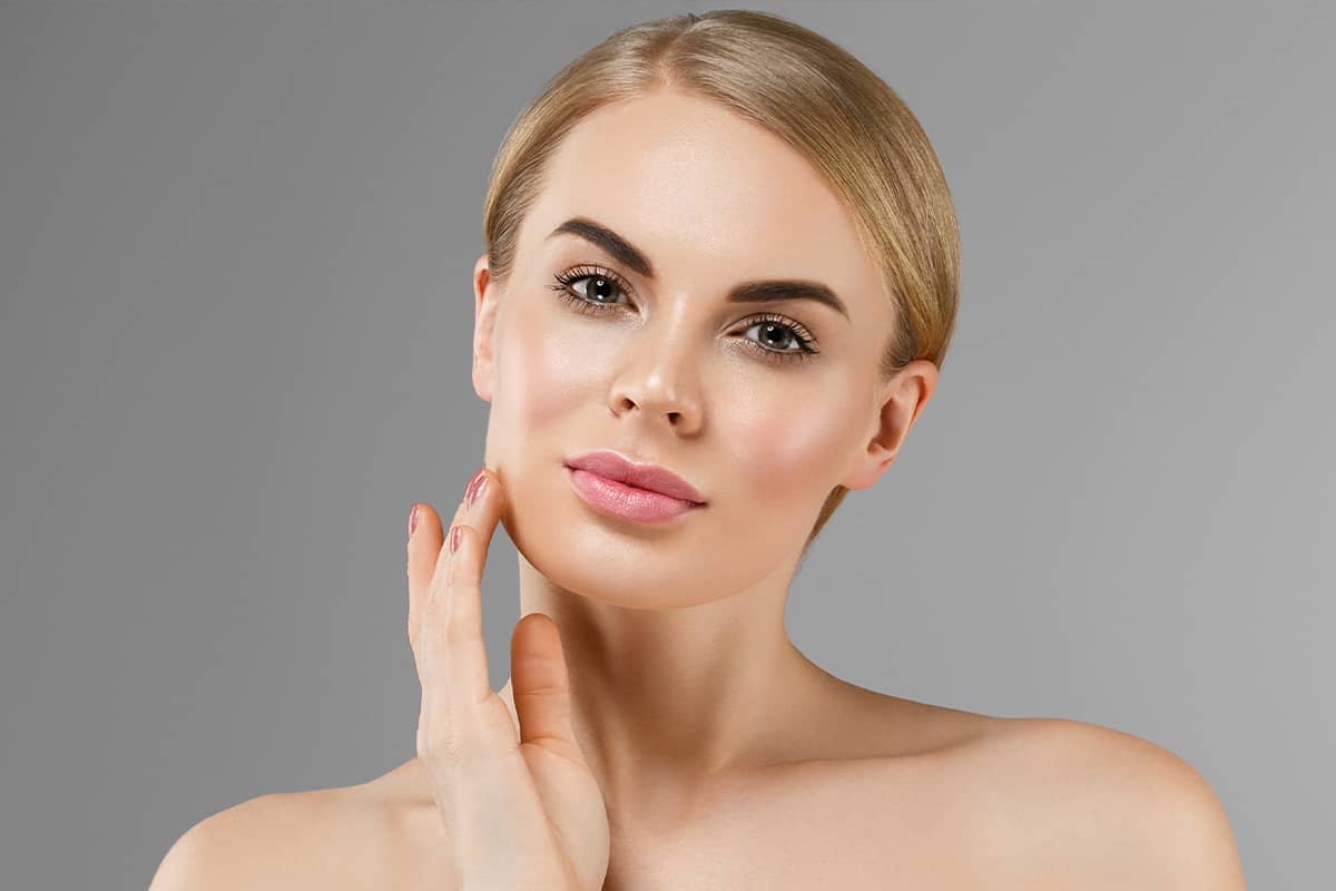 A blonde woman with her hair pulled back looking directly forward while holding the fingertips of one hand against her lower cheek and jawline.