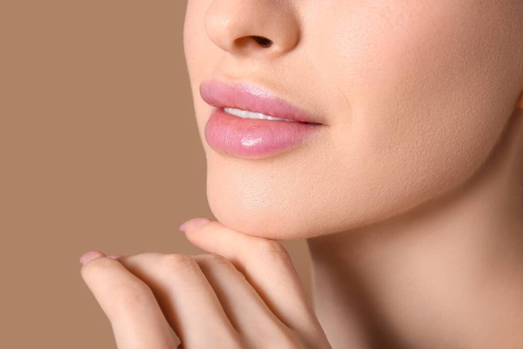 A close-up view of a woman's full, glossy pink lips and lower nose, with her hand gently resting under her chin.